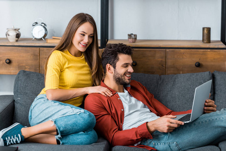 年轻的幸福夫妇坐在沙发上，手提电脑，一边看屏幕一边微笑