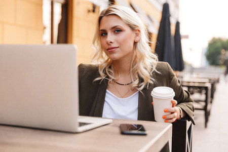 一位年轻漂亮的女士坐在户外咖啡馆里，一边喝外卖咖啡，一边用银色笔记本电脑