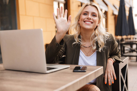 一位身穿夹克衫银色笔记本电脑和智能手机坐在户外咖啡馆的快乐金发女子的肖像