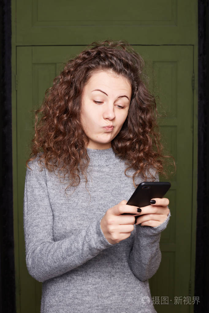 犹豫不决的美国年轻女性手持手机弯着嘴唇有着完美的卷曲健康的头发穿