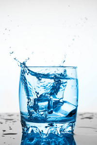 水溅在玻璃杯里，白色背景上有一块冰，一块冰掉进玻璃杯里的水，溅出的水向四面八方飞去