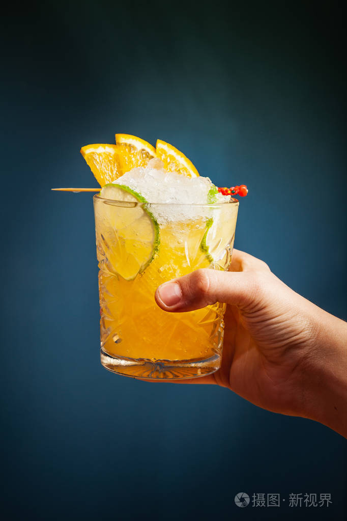 手拿橙色酒精鸡尾酒与冰橙和石灰在趋势蓝色背景酒吧概念视图复制品牌