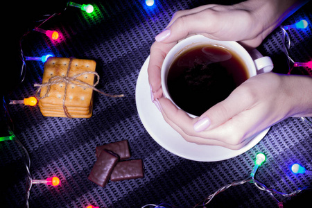 概念新年。 圣诞故事。 女孩把手放在一杯热茶饼干和糖果上，放在一帧圣诞花环上。 黑暗的灯光。 从上面看。 童话氛围和奇迹的迷雾