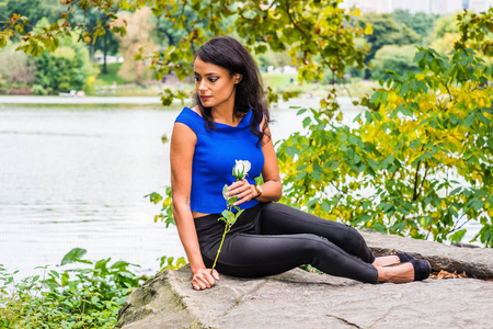 我想你在想你。 年轻的美国女人穿着蓝色无袖衬衫，黑色裤子，手里拿着白色玫瑰花，坐在纽约中央公园湖边绿树下的岩石上。