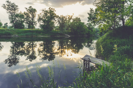 夏天日落时在河上钓鱼的木制平台。 水中树木和天空的倒影。