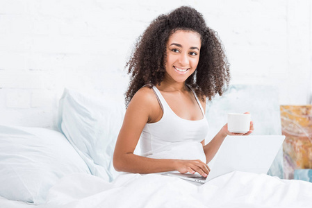 快乐的卷曲女孩拿着咖啡杯，在家里的早晨在床上使用笔记本电脑