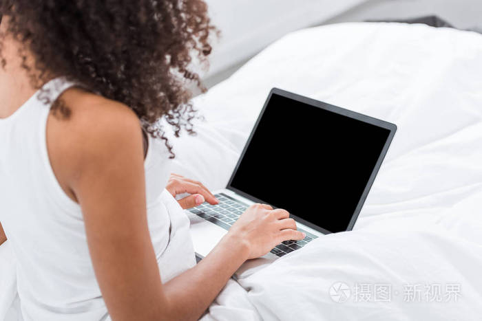 早晨在家的时候在床上使用带有空白屏幕的笔记本电脑的卷曲女孩的局部