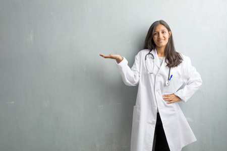 年轻的印度医生女人靠在墙上，手里拿着东西，手里拿着一件东西，展示着一种微笑和快乐的产品，提供了一个想象中的物体