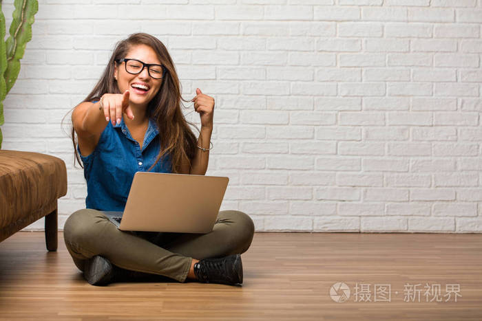 年轻的拉丁女人坐在地板上大喊大叫，嘲笑另一个嘲弄和失控的概念。 拿着笔记本电脑。