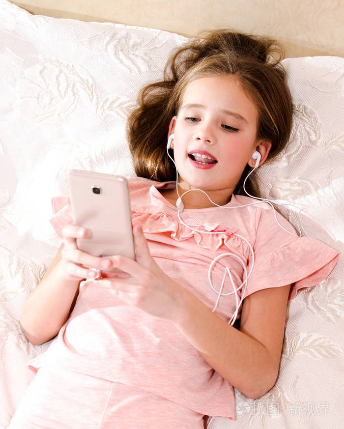 可爱的小女孩躺在床上,用智能手机听音乐唱歌,听耳机耳机