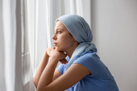 患有卵巢癌的体贴的年轻女孩戴着蓝色头巾，在医疗中心透过窗户看着。