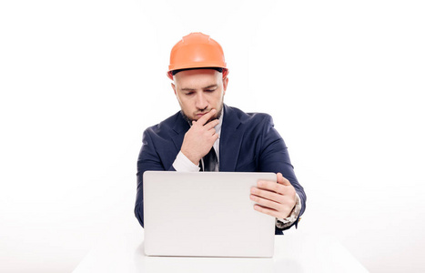 一个穿着橙色头盔的疲惫的建筑商人看着笔记本电脑屏幕，研究建筑项目。坐在桌边喝咖啡，紧张