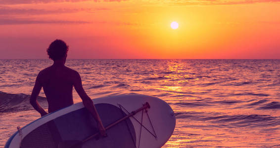 无法辨认的年轻人带着冲浪板在日落时出海。