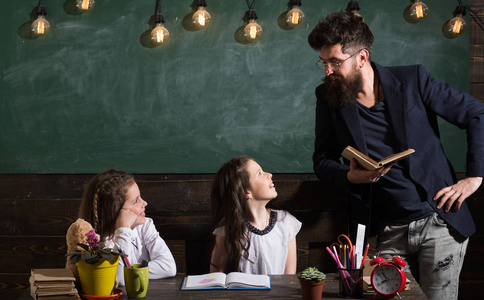 留胡子的男人教女学生看书。好奇开朗的孩子听老师的注意。老师和女孩学生在教室, 黑板上的背景。小学理念