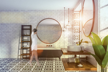 女性在现代浴室的内部，砖墙瓷砖地板，黑色浴缸，圆形镜子和白色圆形水槽在木制架子上。 左边有毛巾的架子。 色调图像模糊