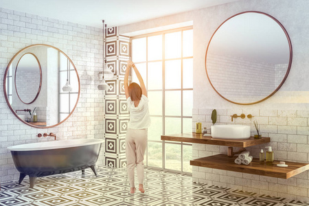 女性在现代浴室的内部，砖墙瓷砖地板，黑色浴缸，圆形镜子和白色圆形水槽在木制架子上。 调色图像