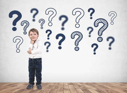 许多问号可爱的金发小男孩穿着白色衬衫,思考着站在混凝土墙附近,上面