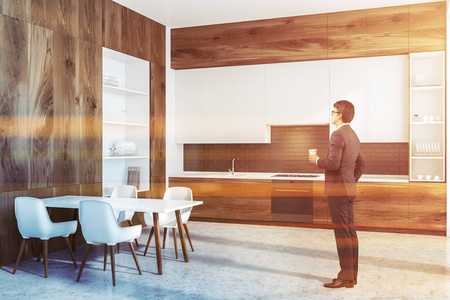 男人在现代厨房的角落，黑色砖墙，混凝土地板，木制台面，白色橱柜，白色和木制橱柜在左边。 带椅子的白色桌子。 调色图像