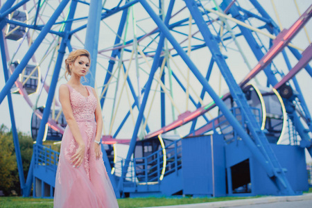 年轻漂亮的女人穿着粉红色的长裙在公园散步。 摩天轮附近户外美丽女孩的时尚风格肖像。