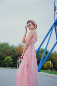 年轻漂亮的女人穿着粉红色的长裙在公园散步。 摩天轮附近户外美丽女孩的时尚风格肖像。