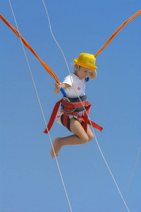 一个小女孩在蹦床上跳得很高，蹦床上用橡胶绳抵着蓝天和白云。 冒险和极限运动。 暑假跳跃的概念。
