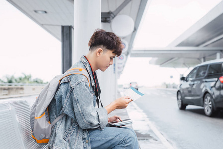 耳机的家伙年轻人坐着等公共汽车. 他拿着背包,听着耳机里的音乐.