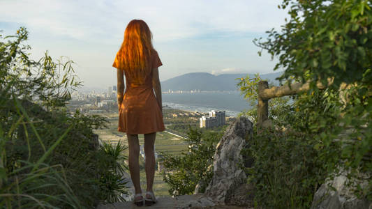 红色头发的旅行女孩正在大理石山上旅行，住在山顶上，看着夕阳下的大南城和越南的朱尼尔西亚和山脉的风景旅行概念