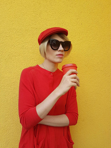 女性模特穿着长红色毛衣和红色帽子, 红色纸杯站在黄色背景上。美丽的高加索妇女在毛衣在咖啡馆前喝咖啡的户外照片