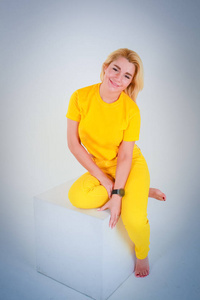 白色至浅蓝色渐变背景..一个穿着黄色衬衫的女孩坐在一个白色的立方体上，微笑着全长。