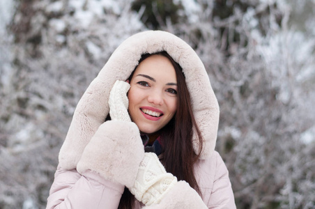 年轻美丽的情感女人的肖像，穿着带帽的羽绒衣和手套，在白雪覆盖的花园背景。冬季雪景