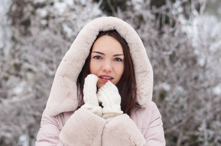 年轻美丽的情感女人的肖像，穿着带帽的羽绒衣和手套，在白雪覆盖的花园背景。冬季雪景