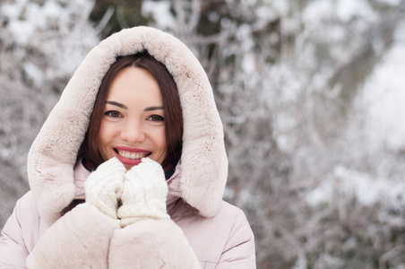 年轻美丽的情感女人的肖像，穿着带帽的羽绒衣和手套，在白雪覆盖的花园背景。冬季雪景