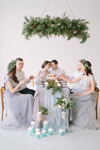 新娘新郎伴娘和小妇人一组人坐在婚礼桌前，手里拿着结婚蛋糕松木装饰和白色装饰的大厅里的蜡烛