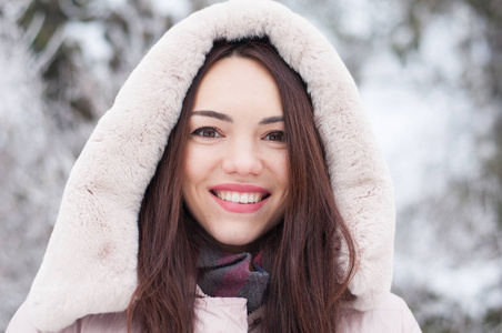 年轻美丽的情感女人的肖像，穿着带帽的羽绒服和手套，在白雪覆盖的花园背景上。 冬季雪景