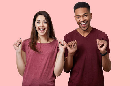 室内镜头快乐的跨种族夫妇紧握拳头，欣喜若狂的面部表情庆祝成功，穿着休闲T恤，孤立于粉红色背景。 多民族关系概念