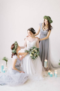 美丽的新娘穿着白色婚纱，手捧花束，坐在老式椅子上，伴娘穿着蓝色灰色连衣裙和花环。 婚礼装饰和背景上的蜡烛