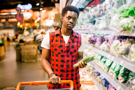 非洲人在超市的蔬菜区购物。 黑人在市场上购物，同时买蔬菜。 帅哥拿着购物篮阅读产品的营养价值。
