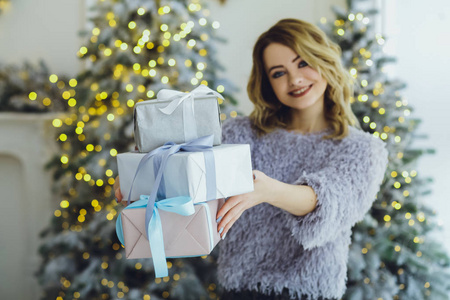 年轻漂亮的金发女郎穿着灰色毛衣，手上拿着礼物，靠近新年圣诞树，带着礼物
