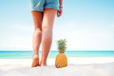 在夏天放松和休闲，晒黑的年轻女人的腿与菠萝站在热带海滩在夏天。 复古色彩色调效果