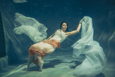 优雅苗条的女孩在水下游泳，就像一个自由潜水员穿着白色的晚礼服和美丽的织物