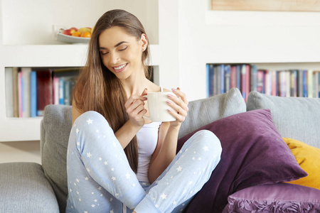 照片中年轻漂亮的女人穿着睡衣，喝着茶，清晨在沙发上放松。
