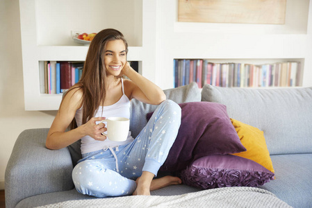 全长镜头迷人的年轻女子穿着睡衣，喝着早茶，在她的公寓沙发上放松。