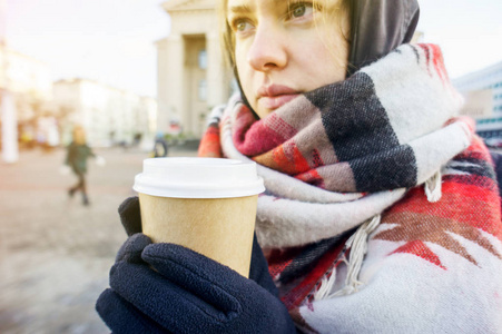 年轻漂亮的女孩戴着手套，戴着一条舒适的大围巾，在城市环境中拿着一杯外卖咖啡。 冬季咖啡的概念