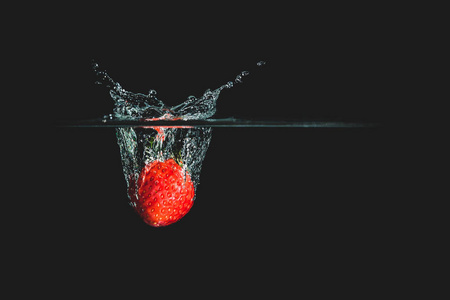 一种新鲜美味的红色草莓，在黑色的背景下分离出来。 红色草莓掉在水里，引起飞溅。 健康饮食消费水果的概念。