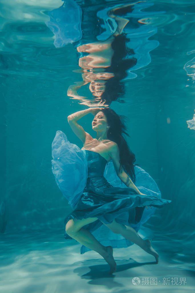美丽的模特女孩,留着长长的黑发,穿着晚蓝色连衣裙在水下游泳,享受
