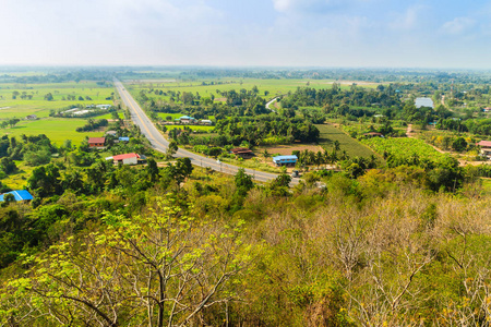 从山顶俯瞰公路，周围有农村绿色稻田和蓝天背景。 在泰国菲希特省，有汽车和卡车穿过山丘的省道。