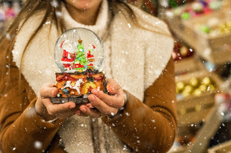 在圣诞博览会上, 穿着暖和外套的女孩在一个商场里拿着玻璃球, 上面有冷杉房子和人造雪。冬季心情