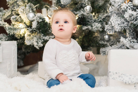 小可爱的女婴穿着白色毛衣，背景是圣诞树灯花环和礼品盒。 新年和圣诞节假期。