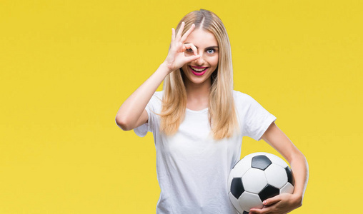 年轻漂亮的金发女郎把足球抱在孤立的背景上，脸上带着快乐的笑容，做着好的标志，用手盯着手指看