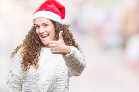年轻的黑发女孩戴着圣诞帽在孤立的背景上，用手做快乐的竖起大拇指的手势。 赞许的表情看着相机，显示出成功。